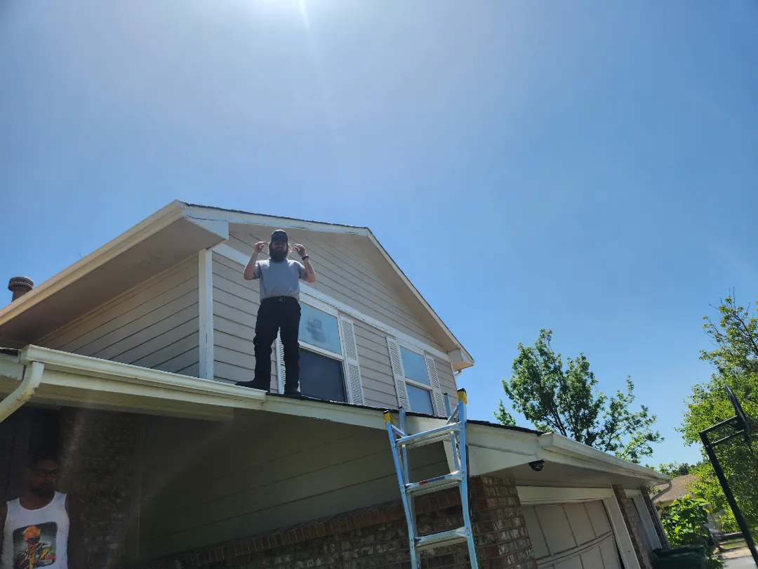 A person stands on the edge of a house rooftop, holding materials, with a ladder leaning against the side and a clear blue sky above.