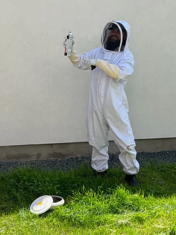 A person in a white protective suit and gloves holds a canister while standing on grass near a white wall.